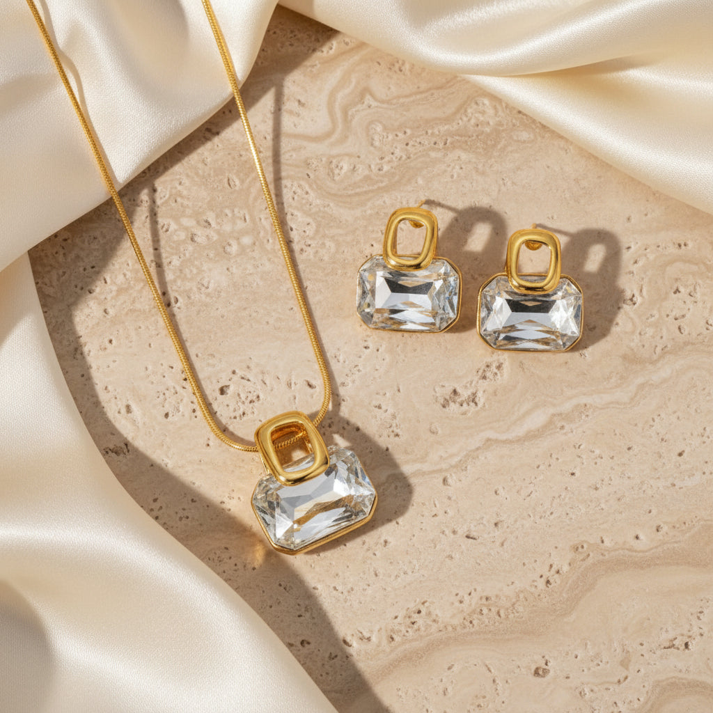 Gold jewelry set featuring a pendant necklace and two matching stud earrings with clear rectangular faceted gemstones, displayed on a gray surface with white blocks.