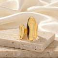 A minimalist gold ring with a large, organic-shaped face and a smaller matching accent piece, displayed on a white curved surface.