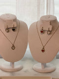 Two mannequin heads with necklaces with heart-shaped pendants and matching earrings on a neutral background.