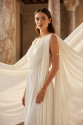 A woman wearing a flowing draped white gown with gold bangles, standing in a classical interior with marble columns and draped fabric.