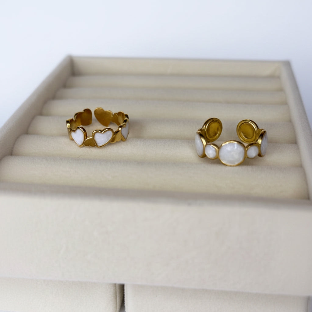 Two Gold and white pearl open rings on a beige cushion with a white and gold background. One ring has heart shapes and the other is circles