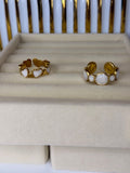 Two Gold and white pearl open rings on a beige cushion with a white and gold background. One ring has heart shapes and the other is circles