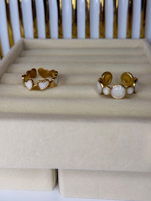 Two Gold and white pearl open rings on a beige cushion with a white and gold background. One ring has heart shapes and the other is circles