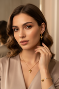 A close-up lifestyle shot of a woman with wavy brown hair wearing a gold heart pendant necklace, matching gold heart earring, and a delicate gold bracelet with a small white stone, set against a warm beige background.