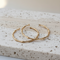 A pair of gold-toned cuffs with a bamboo branch design, resting on a textured beige stone slab.