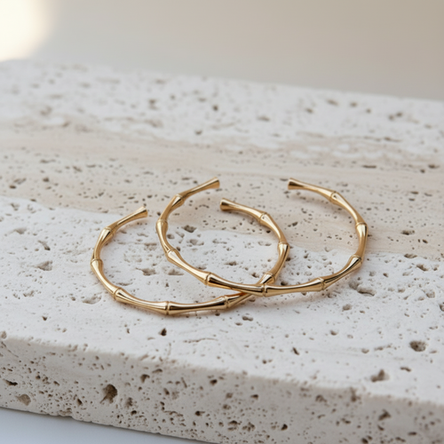 A pair of gold-toned cuffs with a bamboo branch design, resting on a textured beige stone slab.