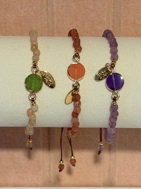 Three beaded bracelets with colorful charms on a white cylindrical stand against a beige background.