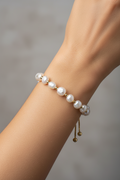 A close-up of a person's wrist wearing a delicate bracelet made of white freshwater pearls separated by small gold spacer beads, with a gold clasp and dangling bead charm.
