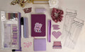 Collection of purple stationery items including a notebook, pens, and decorative elements on a light surface.