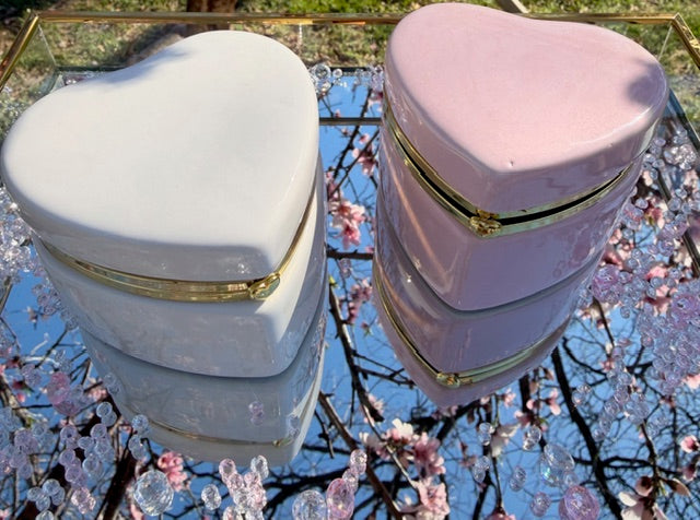 Two heart-shaped boxes, one white and one pink, with gold accents on a reflective surface with cherry blossoms.