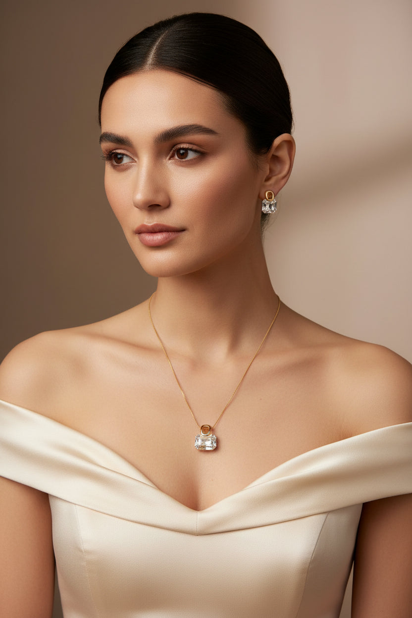 A gold-tone jewelry set featuring a pendant necklace and matching stud earrings displayed on a beige fabric jewelry bust. The pieces have clear faceted stones set in gold-toned metal.