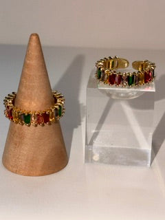 A gold adjustable rings with alternating red and green rectangular gemstones, displayed on a wooden cone stand and a clear acrylic block against a light gray background.