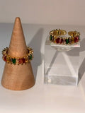A gold adjustable rings with alternating red and green rectangular gemstones, displayed on a wooden cone stand and a clear acrylic block against a light gray background.