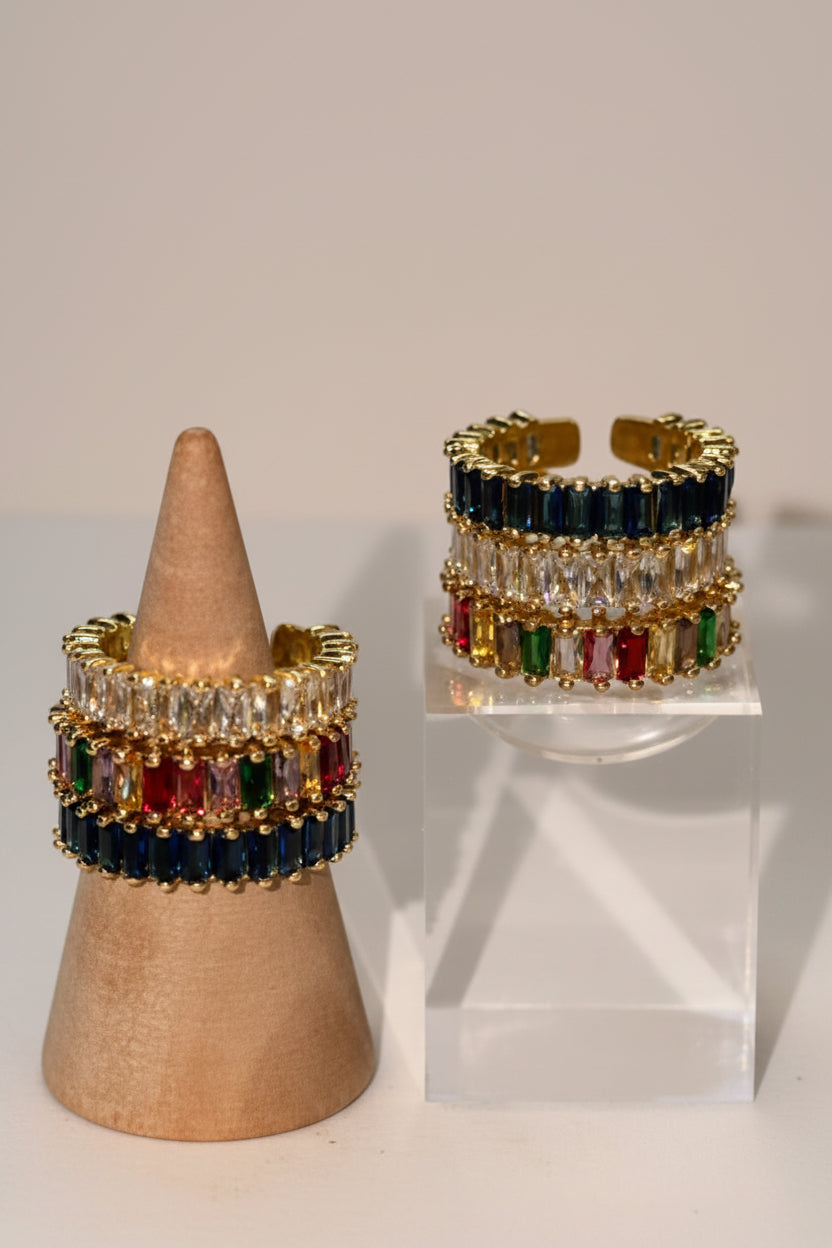 A pair of gold-toned adjustable rings with rows of dark blue, clear, red, and green stones, displayed on a wooden cone stand and a clear acrylic riser against a light gray background.